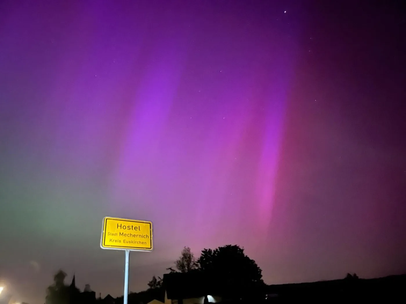 Dasselbe Ortsschild in der Nacht, darüber leuchtet der Himmel in Violett und Grün