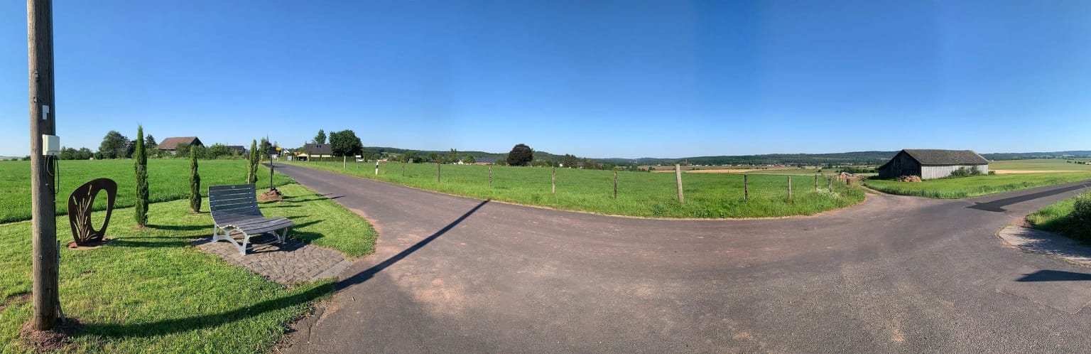 Weiter Blick vom höchsten Punkt des Dorfes über Wiesen und Felder, vorne eine geschwungene Bank