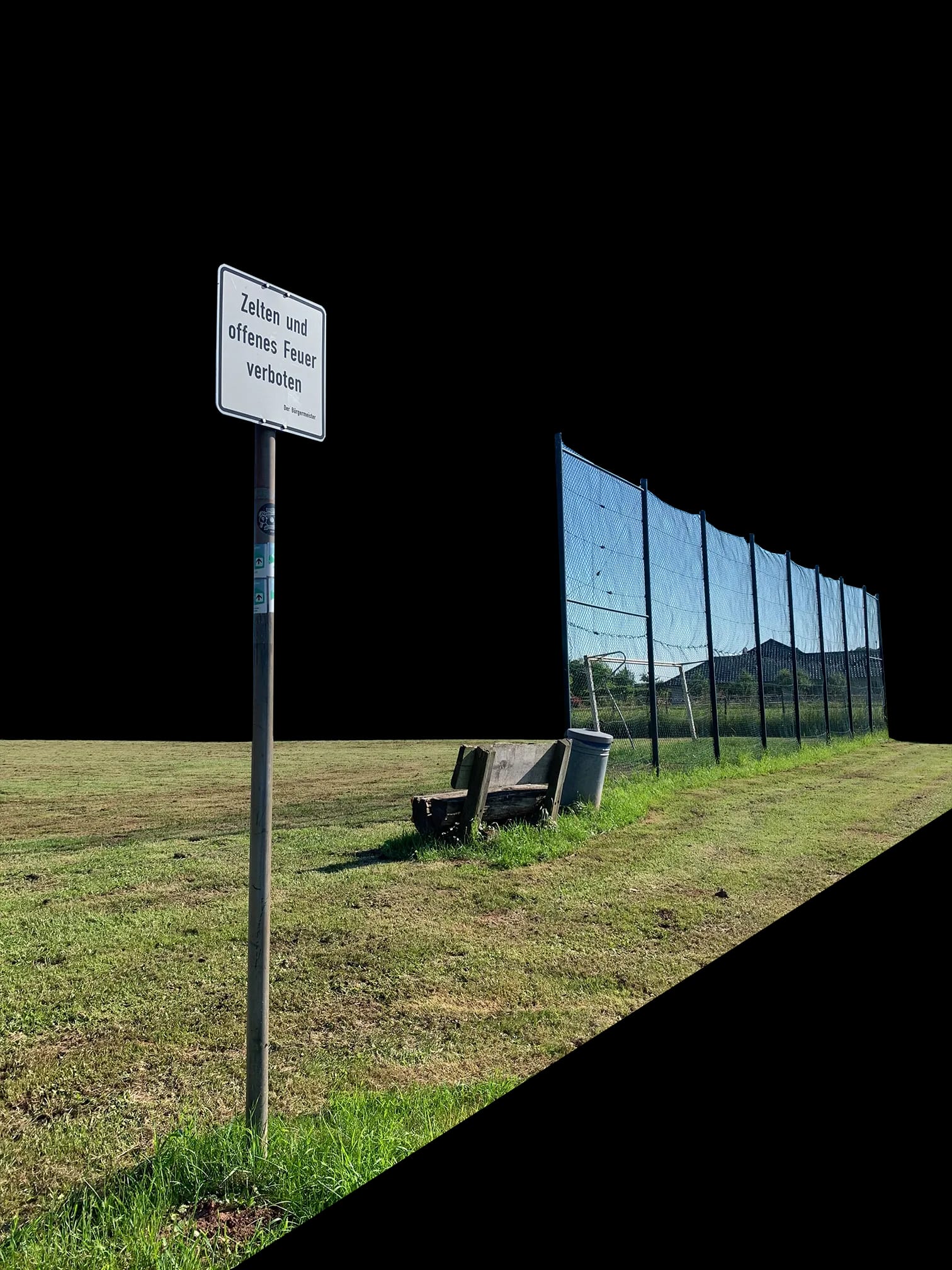 Der Bolzplatz mit Hinweisschild, einer Bank und dem hohen Ballfangzaun am Rand