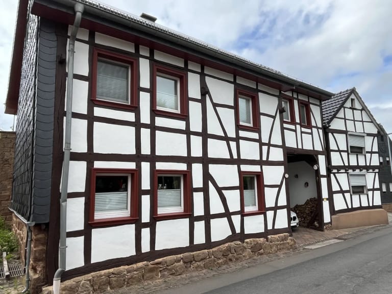 Weißes Fachwerkhaus mit dunkelrotem Gebälk und Sockel aus rotem Sandstein an einer ansteigenden Straße