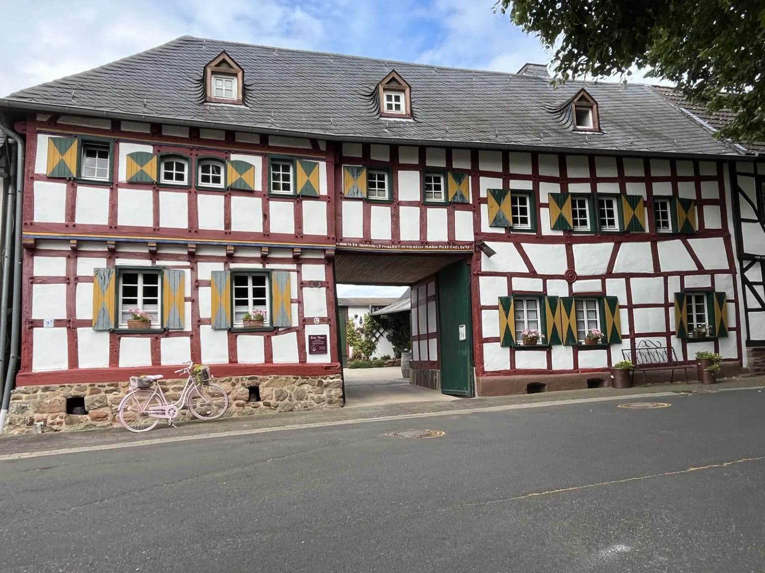 Langes weißes Fachwerkhaus mit rotem Gebälk, gelb-grünen Fensterläden und einer Toreinfahrt in der Mitte