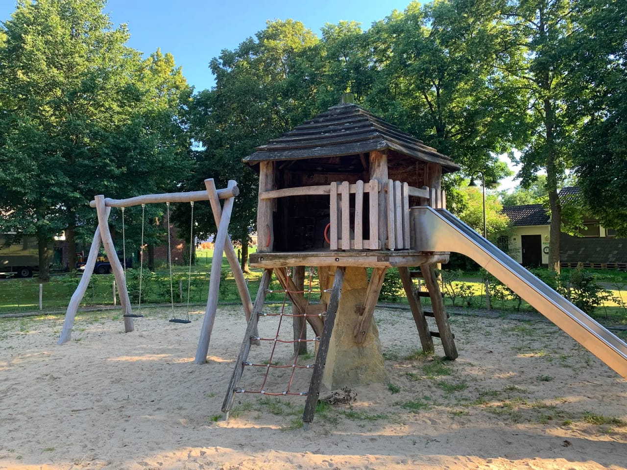Der Spielplatz unter Laubbäumen: ein Holzhaus auf Stelzen mit Rutsche, daneben ein Schaukelgerüst, darunter eine Sandfläche