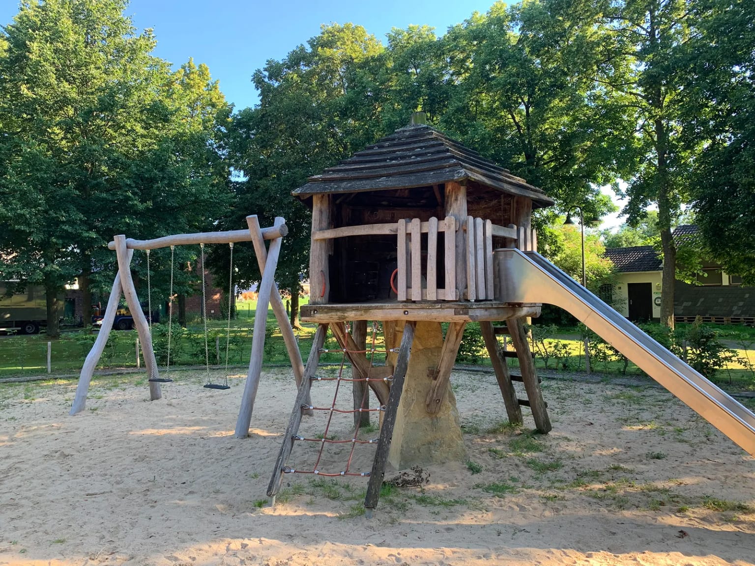 Der Spielplatz unter Laubbäumen: ein Holzhaus auf Stelzen mit Rutsche, daneben ein Schaukelgerüst, darunter eine Sandfläche