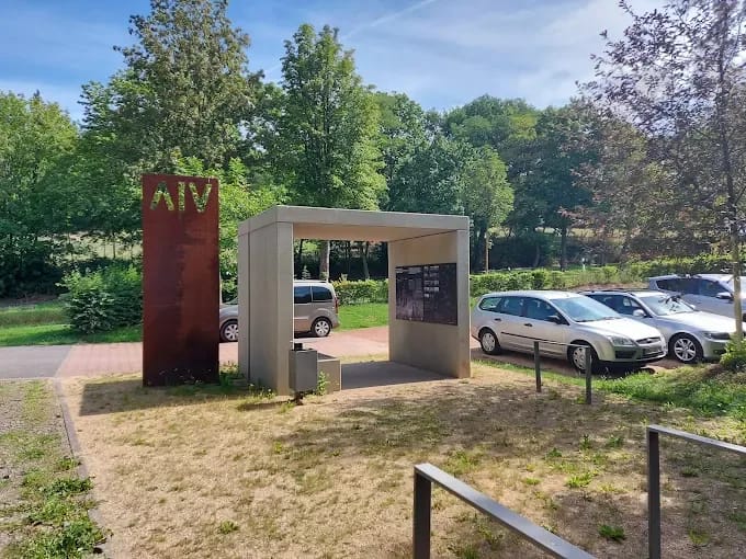 Rastplatz Mansio: ein offener Pavillon aus hellem Beton neben einer rostroten Stele, dahinter der Parkplatz unter Bäumen