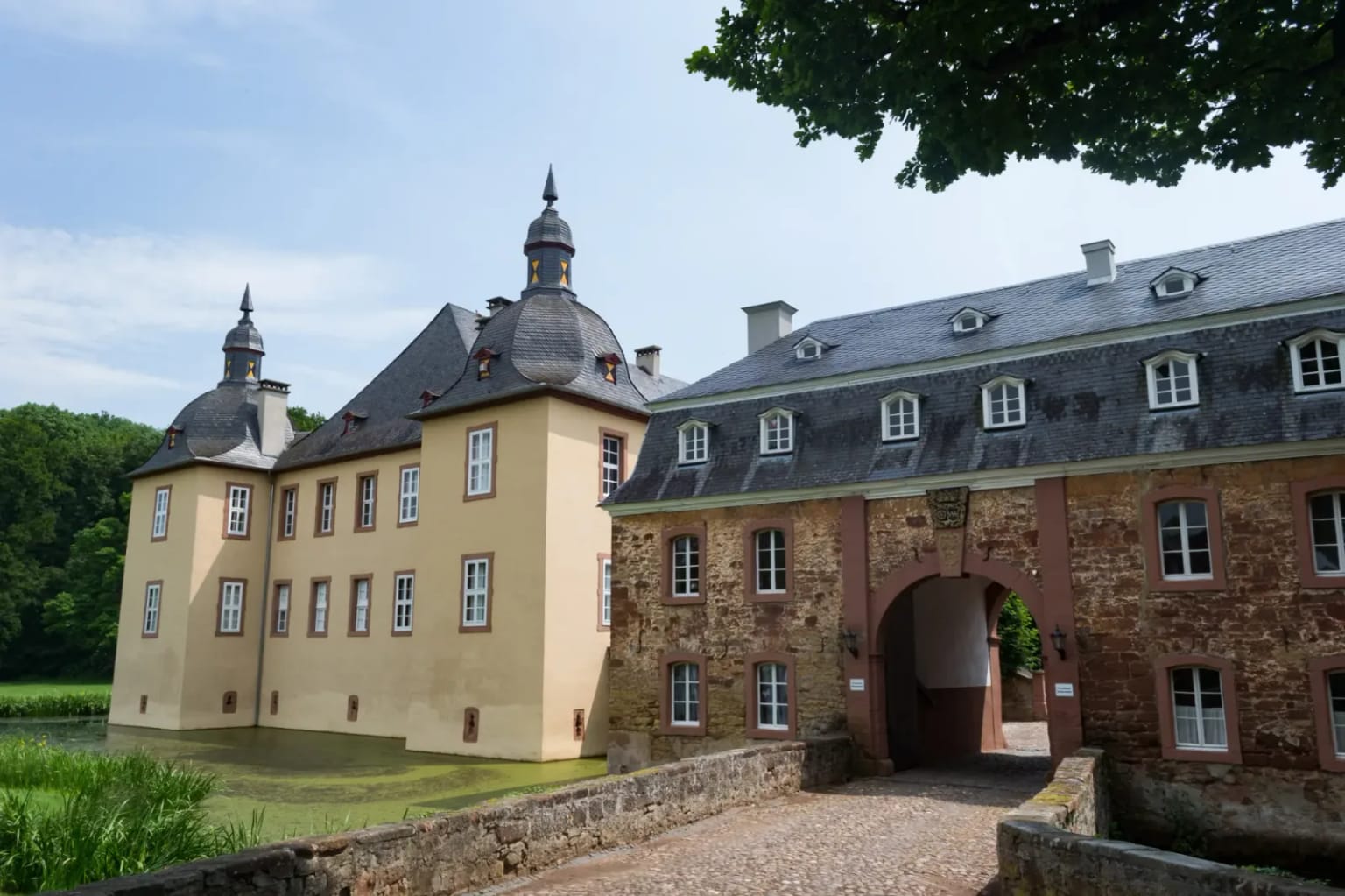 Schloss Eicks: hell verputzte Wasserburg mit Schieferdächern und Türmen, davor ein Wassergraben und eine Brücke zum Tor